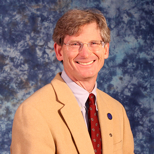 Man wearing a tan suit and maroon tie in front of a blue background
